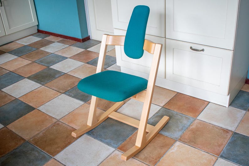 May include: A teal ergonomic chair with a wooden frame. The chair has a curved backrest and seat, and a rocking base. The chair is set against a white cabinet and a tiled floor.