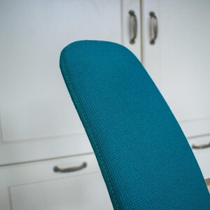 May include: Close-up of a teal-colored chair back with a textured fabric. The chair back is curved and set against a white cabinet with dark handles. The chair is part of a furniture set.