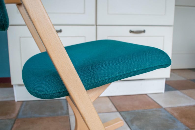 May include: A teal ergonomic kneeling chair with a wooden frame. The seat and knee rest are upholstered in a textured teal fabric. The chair's design promotes good posture and is suitable for home or office use.