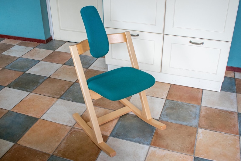 May include: A teal kneeling chair with a wooden frame. The chair has a padded seat and backrest, and a rocking base. The chair is designed to promote good posture and is suitable for use at a desk or table.