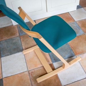 May include: A teal-colored chair with a wooden frame. The chair has a curved back and seat, and the frame is made of light-colored wood. The chair is positioned on a tiled floor with various earth-toned squares.