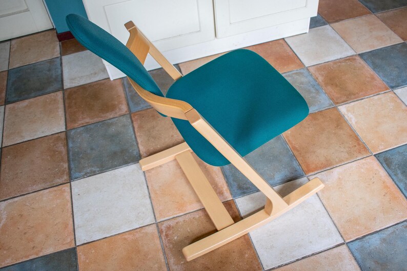 May include: A teal-colored chair with a wooden frame. The chair has a curved back and seat, and the frame is made of light-colored wood. The chair is positioned on a tiled floor with various earth-toned squares.