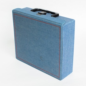 May include: A rectangular blue denim-textured carrying case featuring a prominent red stitched border along the front panel. The case includes a black plastic handle attached to the top edge, providing a functional grip for transport and storage.
