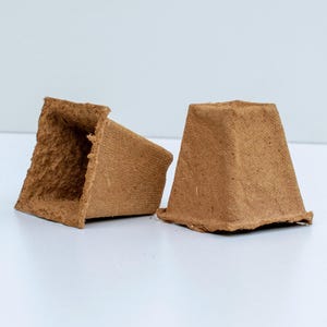 Op de afbeelding: Twee kleine, bruine, vierkante, biologisch afbreekbare plantenpotten. Eén pot ligt op zijn kant, de andere staat rechtop. De potten zijn gemaakt van een getextureerd, vezelachtig materiaal, waarschijnlijk voor het planten van zaailingen.