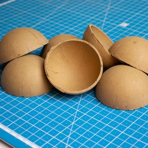 Op de afbeelding: Een verzameling onafgewerkte, lichtbruine papieren mache halve bollen. De open helften liggen op een blauwe snijmat met een wit rasterpatroon. Deze knutselbenodigdheden zijn ideaal voor doe-het-zelf projecten.