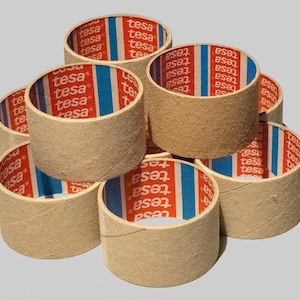 Puede incluir: Un conjunto de rollos cilíndricos de cinta adhesiva de cartón. Los rollos son de color beige y muestran la marca "tesa" en su interior en rojo, blanco y azul. Están apilados en forma de pirámide.