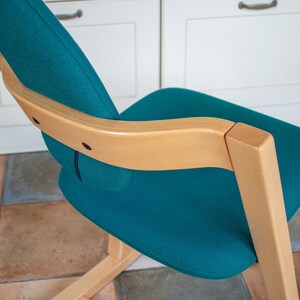 May include: Teal fabric chair with a light wood frame. The chair has a curved back and seat, with a natural wood armrest. The chair is set against a white cabinet and a tiled floor.