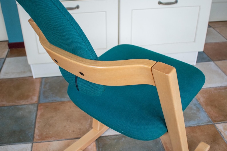 May include: Teal fabric chair with a light wood frame. The chair has a curved back and seat, with a natural wood armrest. The chair is set against a white cabinet and a tiled floor.