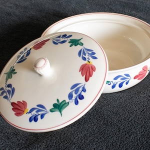 Op de afbeelding: Een witte keramische ovenschaal met deksel, versierd met een bloemmotief in rood, blauw en groen. De schaal heeft een handvat en een bijpassend deksel met een knop. Het ontwerp herhaalt zich rond de rand van de schaal en het deksel.