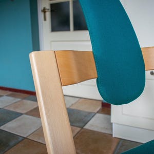 May include: A wooden chair with a teal-colored padded backrest and seat. The chair has a light-colored wooden frame and is set against a backdrop of a blue wall and checkered floor.