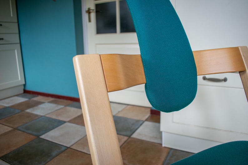 May include: A wooden chair with a teal-colored padded backrest and seat. The chair has a light-colored wooden frame and is set against a backdrop of a blue wall and checkered floor.