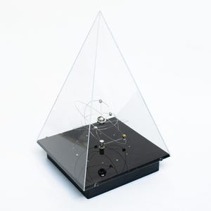 Puede incluir: Escultura cinética negra y transparente, con cubierta piramidal de acrílico. En el interior, esferas y alambres metálicos simulan órbitas planetarias. La base es una plataforma rectangular negra.