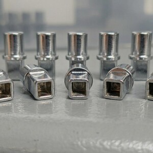 May include: A collection of shiny, silver lug nuts. The lug nuts have a square head and a cylindrical body. The image shows the lug nuts arranged on a gray surface, with some standing upright and others lying on their sides.