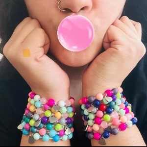 May include: A person wearing a stack of colorful beaded bracelets with silver heart charms on each wrist.