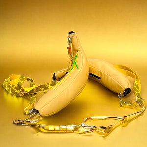 Peut inclure: Deux sacs jaunes en forme de banane avec des sangles jaunes et vertes. Les sacs sont posés sur une surface dorée.