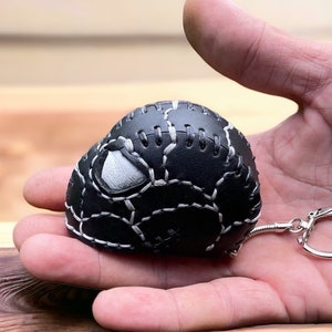 May include: A black leather keychain shaped like a baseball, with white stitching and a white accent. The keychain has a silver clasp and chain. The baseball-shaped item is held in a person's hand.