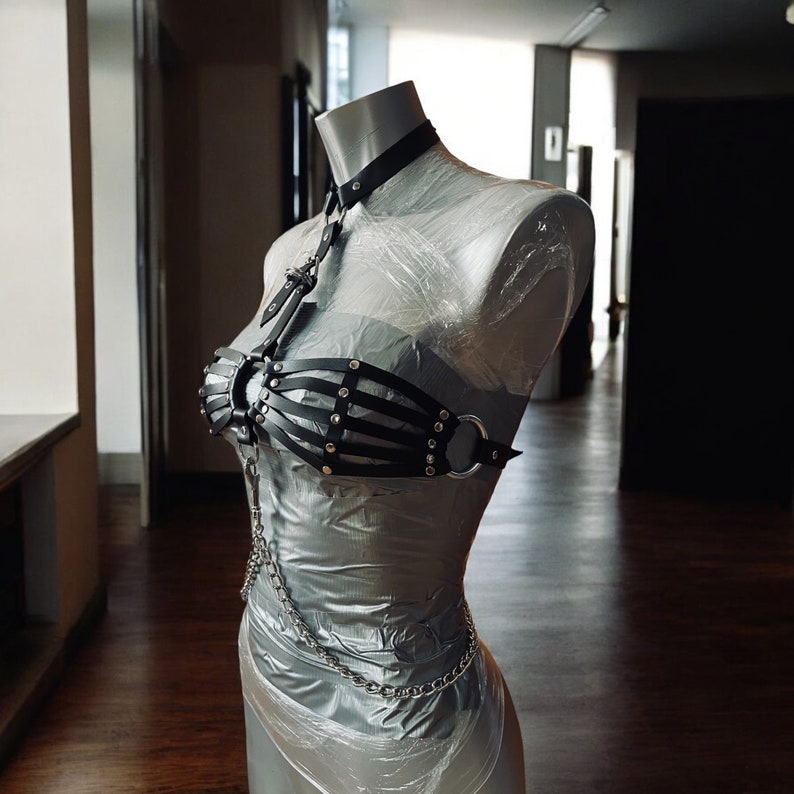 May include: A black leather harness with silver hardware and a chain detail. The harness is on a mannequin and has a bustier style top.