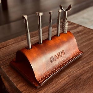 K&ouml;nnte beinhalten: Ein brauner Leder-Werkzeughalter mit dem gepr&auml;gten Wort "GAIUS". Er enth&auml;lt f&uuml;nf Metallwerkzeuge unterschiedlicher Formen und Gr&ouml;&szlig;en. Der Halter steht auf einer Holzoberfl&auml;che.