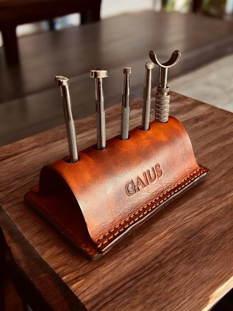 K&ouml;nnte beinhalten: Ein brauner Leder-Werkzeughalter mit dem gepr&auml;gten Wort "GAIUS". Er enth&auml;lt f&uuml;nf Metallwerkzeuge unterschiedlicher Formen und Gr&ouml;&szlig;en. Der Halter steht auf einer Holzoberfl&auml;che.
