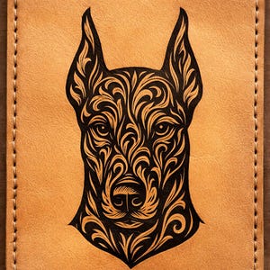 Puede incluir: Artículo de cuero color canela con un diseño floral negro que representa la cara de un perro. La cara del perro está centrada, con orejas largas y patrones detallados. El artículo tiene un borde cosido.