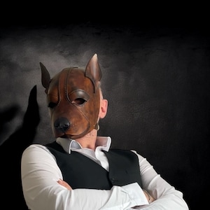 May include: A brown leather dog mask with detailed features, including a snout, ears, and eye openings. The mask is worn over the face, with a black background. The mask is a unique accessory.