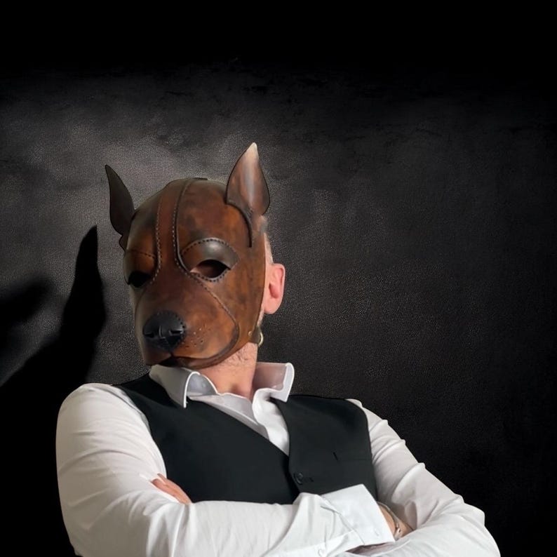 May include: A brown leather dog mask with detailed features, including a snout, ears, and eye openings. The mask is worn over the face, with a black background. The mask is a unique accessory.