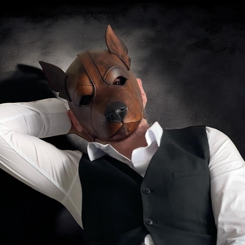 May include: A brown leather dog mask with detailed stitching and eye openings. The mask has a realistic dog snout and ears. The person is wearing a white dress shirt and a black waistcoat. The background is dark.