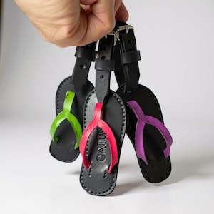 Op de afbeelding: Zwarte leren bagagelabels in de vorm van slippers, met groene, rode en paarse bandjes. Elk label heeft een zwart bandje en een gesp. Het woord "GAIJU" is zichtbaar op het rode label.