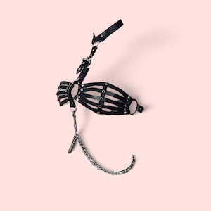 May include: Black leather harness with a chain detail. The harness has a cage-like design with metal studs and a ring on the side.