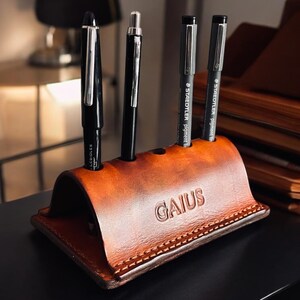 K&ouml;nnte beinhalten: Ein brauner Leder-Stifthalter mit dem gepr&auml;gten Namen "GAIUS". Er enth&auml;lt vier Stifte, zwei schwarz und zwei grau. Der Stifthalter steht auf einer schwarzen Oberfl&auml;che. Das Leder ist braun.