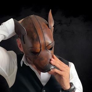 May include: A brown leather dog mask with stitched detailing, featuring pointed ears and a black nose. The mask is worn over the face, with the wearer's hands visible. A unique accessory.