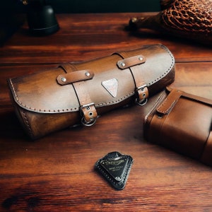 May include: A brown leather motorcycle saddlebag with a metal buckle and a silver Triumph logo. The bag is on a wooden surface with a small metal tag that says "Triumph, Made Under Patent".