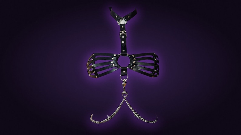 May include: Black leather harness with silver hardware and a chain detail. The harness has a cage-like design and a large ring in the center.