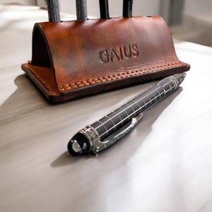 K&ouml;nnte beinhalten: Ein brauner Leder-Schreibtisch-Organizer mit dem gepr&auml;gten Namen "GAIUS" auf der Vorderseite. Der Organizer enth&auml;lt mehrere schwarze Stifte. Ein schwarzer und silberner Stift liegt auf der Oberfl&auml;che vor dem Organizer.