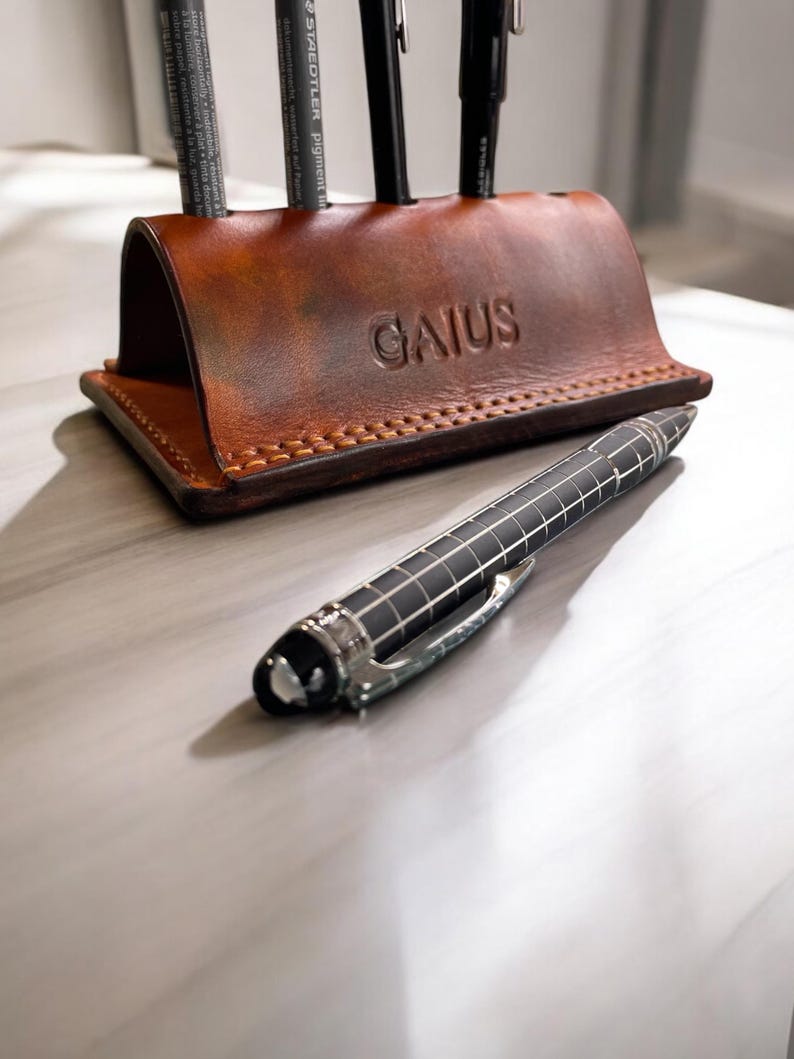 K&ouml;nnte beinhalten: Ein brauner Leder-Schreibtisch-Organizer mit dem gepr&auml;gten Namen "GAIUS" auf der Vorderseite. Der Organizer enth&auml;lt mehrere schwarze Stifte. Ein schwarzer und silberner Stift liegt auf der Oberfl&auml;che vor dem Organizer.