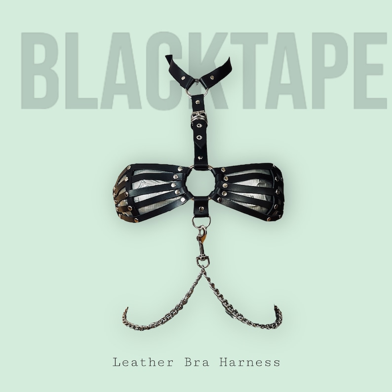 May include: Black leather bra harness with silver hardware and a chain detail. The harness has a choker style neckline and a large O-ring in the center. The text "BLACKTAPE" is displayed above the harness.