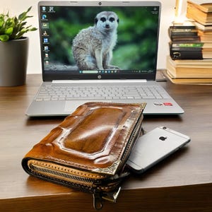 Peut inclure: Un portefeuille en cuir marron avec une fermeture éclair et des coins métalliques décoratifs est posé sur un bureau en bois à côté d'un ordinateur portable argenté et d'un smartphone. L'écran de l'ordinateur affiche un suricate.