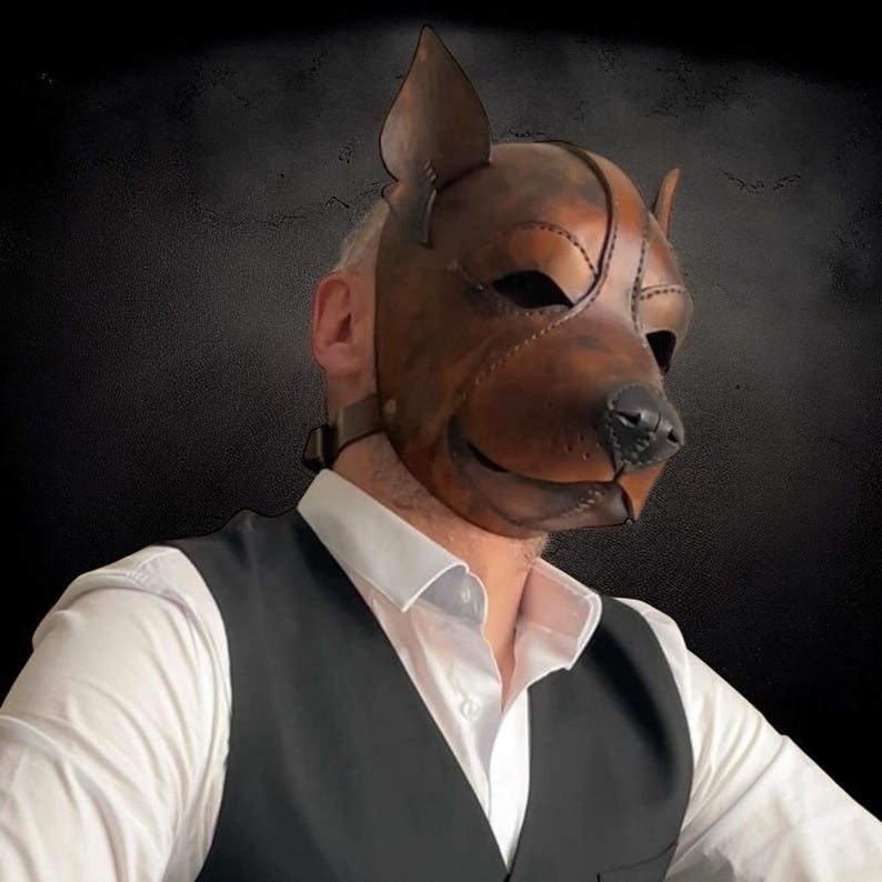 May include: A brown leather dog mask with stitched details, featuring pointed ears and a black nose. The mask is worn with a black waistcoat and white collared shirt. The mask covers the entire face.