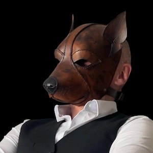 May include: A brown leather dog mask with pointed ears and a black nose. The mask covers the upper face and is secured with a strap. The person is wearing a white dress shirt and a black waistcoat.