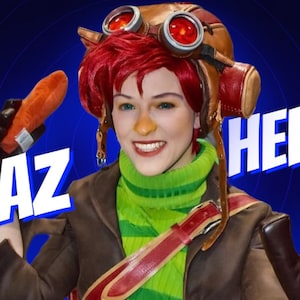 May include: A woman wearing a brown jacket and green sweater with a red and brown aviator helmet with goggles. The helmet has the word "HELMET" written on it. She is holding a plush orange carrot in her right hand and is smiling. The text "RAZ" is written in white letters on a blue background.
