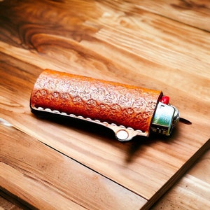 Op de afbeelding: Een bruine leren aanstekerhoes met een zilveren aansteker erin. De hoes heeft een getextureerd oppervlak en een zilveren drukknoop sluiting.