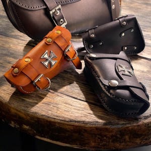 Peut inclure: Sacs de moto en cuir marron et noir, avec des détails argentés et un emblème de croix. Les sacs sont de différentes tailles et sont fermés par des sangles et des boucles.