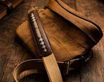 Peut inclure: Un sac messager en toile marron doté d'un rabat avec une bordure en cuir marron foncé. La bandoulière réglable est en tissu tissé beige et comprend une section rembourrée avec un motif à carreaux noir, blanc et beige.
