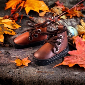 Puede incluir: Un par de botas de cuero marrón con cordones y costuras negras, unidas a un llavero plateado. Las botas están sobre un lecho de hojas de otoño.