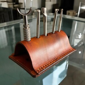 K&ouml;nnte beinhalten: Ein brauner Lederwerkzeughalter f&uuml;r Lederhandwerk. Er enth&auml;lt f&uuml;nf Metallwerkzeuge unterschiedlicher Formen, darunter ein Werkzeug mit gebogener Kante. Der Halter ist f&uuml;r die Lederverarbeitung konzipiert.