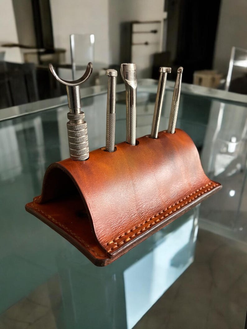 K&ouml;nnte beinhalten: Ein brauner Lederwerkzeughalter f&uuml;r Lederhandwerk. Er enth&auml;lt f&uuml;nf Metallwerkzeuge unterschiedlicher Formen, darunter ein Werkzeug mit gebogener Kante. Der Halter ist f&uuml;r die Lederverarbeitung konzipiert.