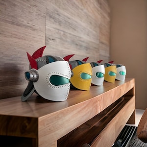 Puede incluir: Cinco cascos de robot de cuero coloridos con una visera turquesa, alineados en un estante de madera. Los cascos son en tonos de blanco, amarillo y gris.