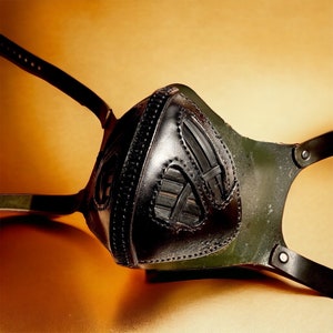 May include: A black leather face mask with a geometric pattern. The mask has a strap that goes around the back of the head.