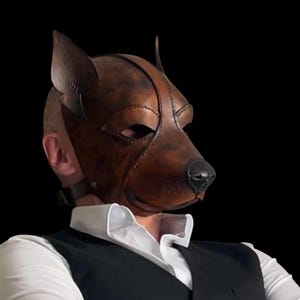 May include: A brown leather dog mask with a realistic design, featuring pointed ears and a black nose. The mask covers the entire face and is secured with a strap. The mask is worn over a white collared shirt and black vest.