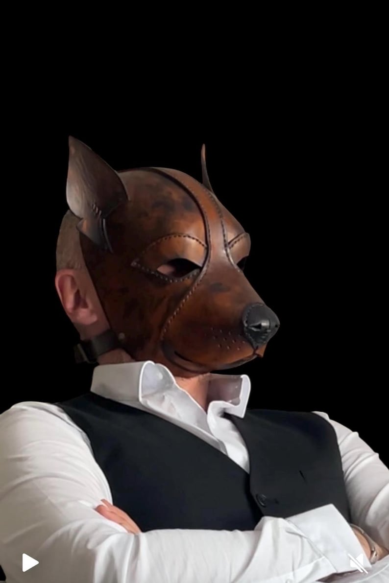 May include: A brown leather dog mask with a realistic design, featuring pointed ears and a black nose. The mask covers the entire face and is secured with a strap. The mask is worn over a white collared shirt and black vest.
